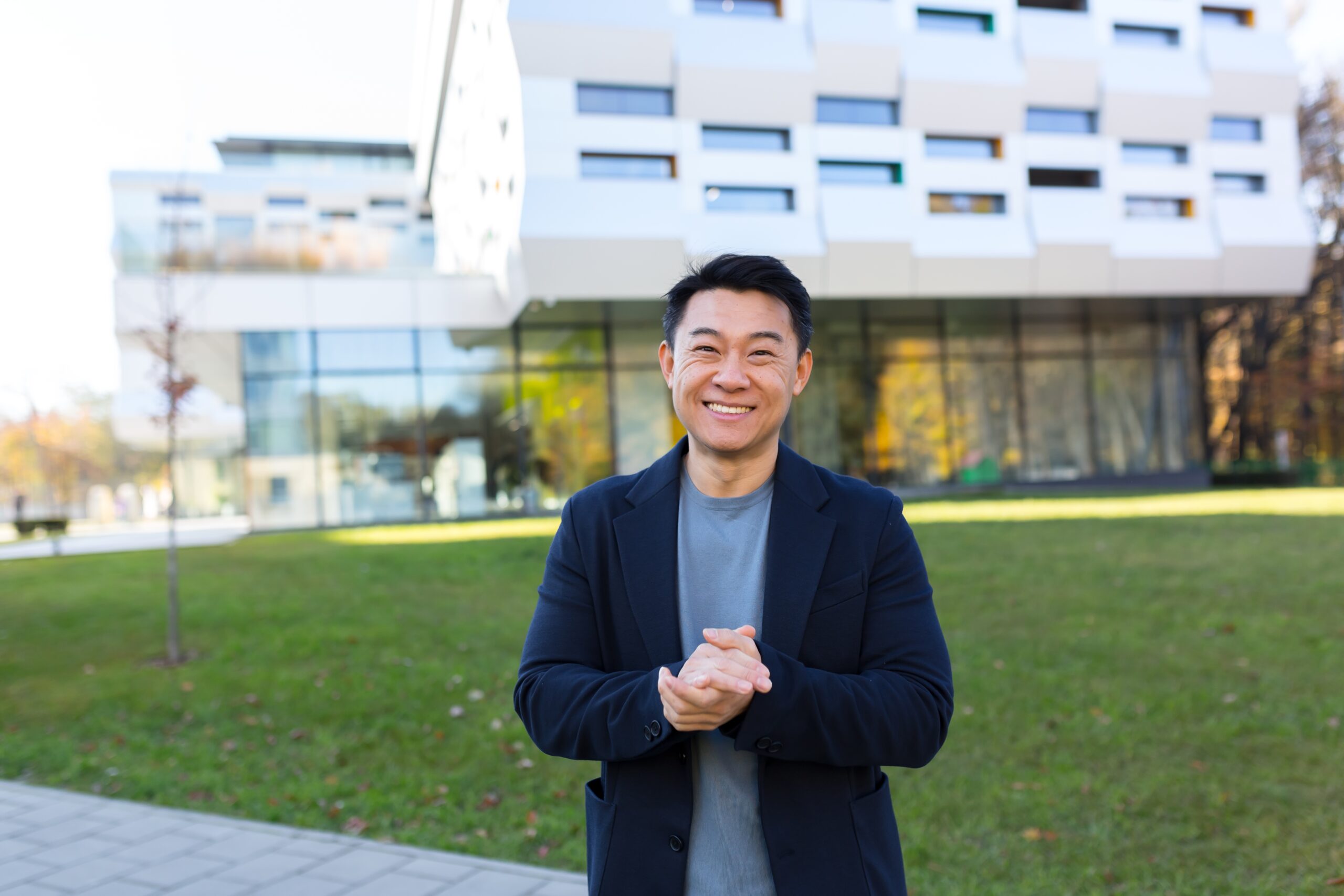 An Asian man stands and smiles with his hands clasped in front of a building.