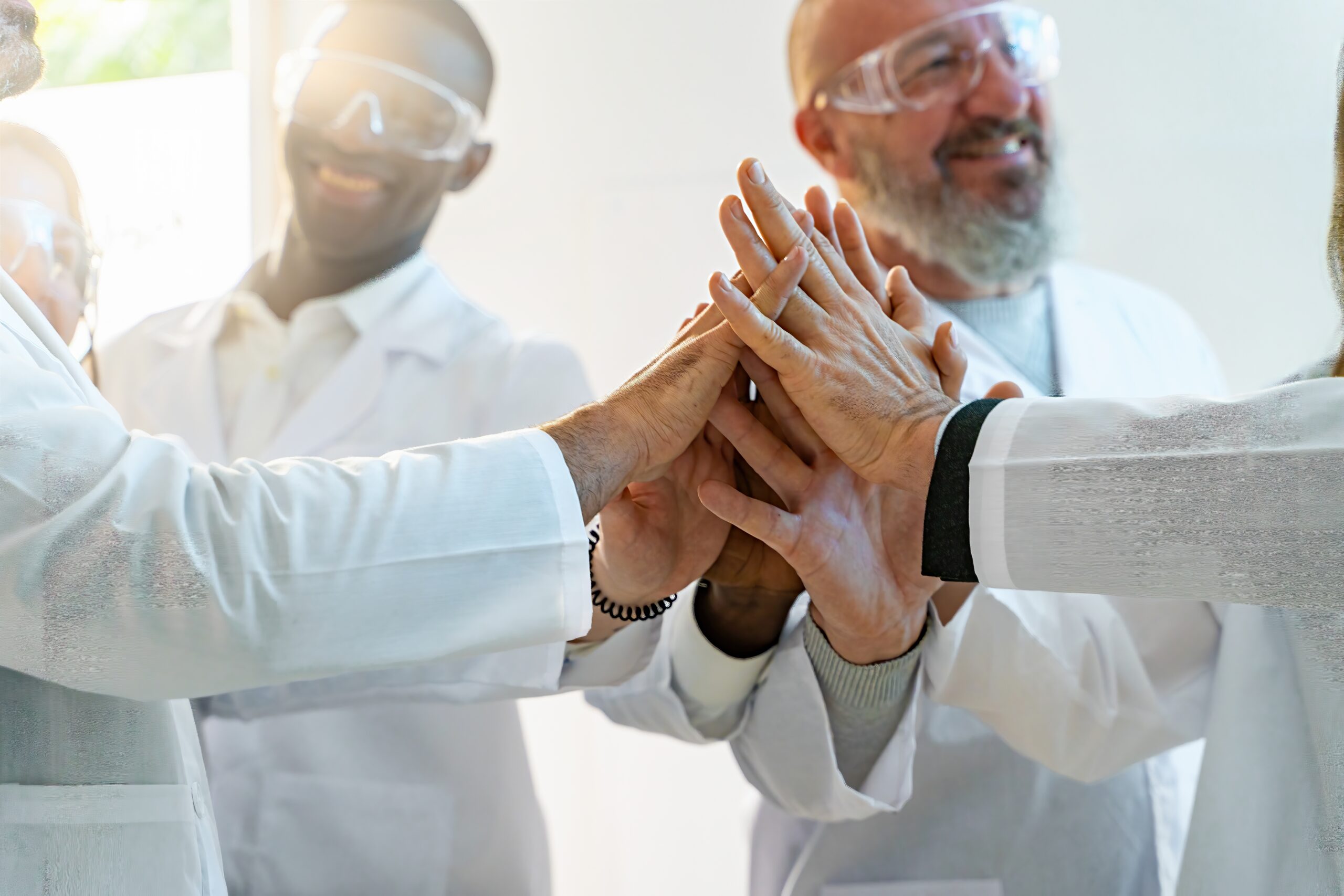 Two male scientists smile and extend their hands to join a collective high five with five other offscreen individuals.