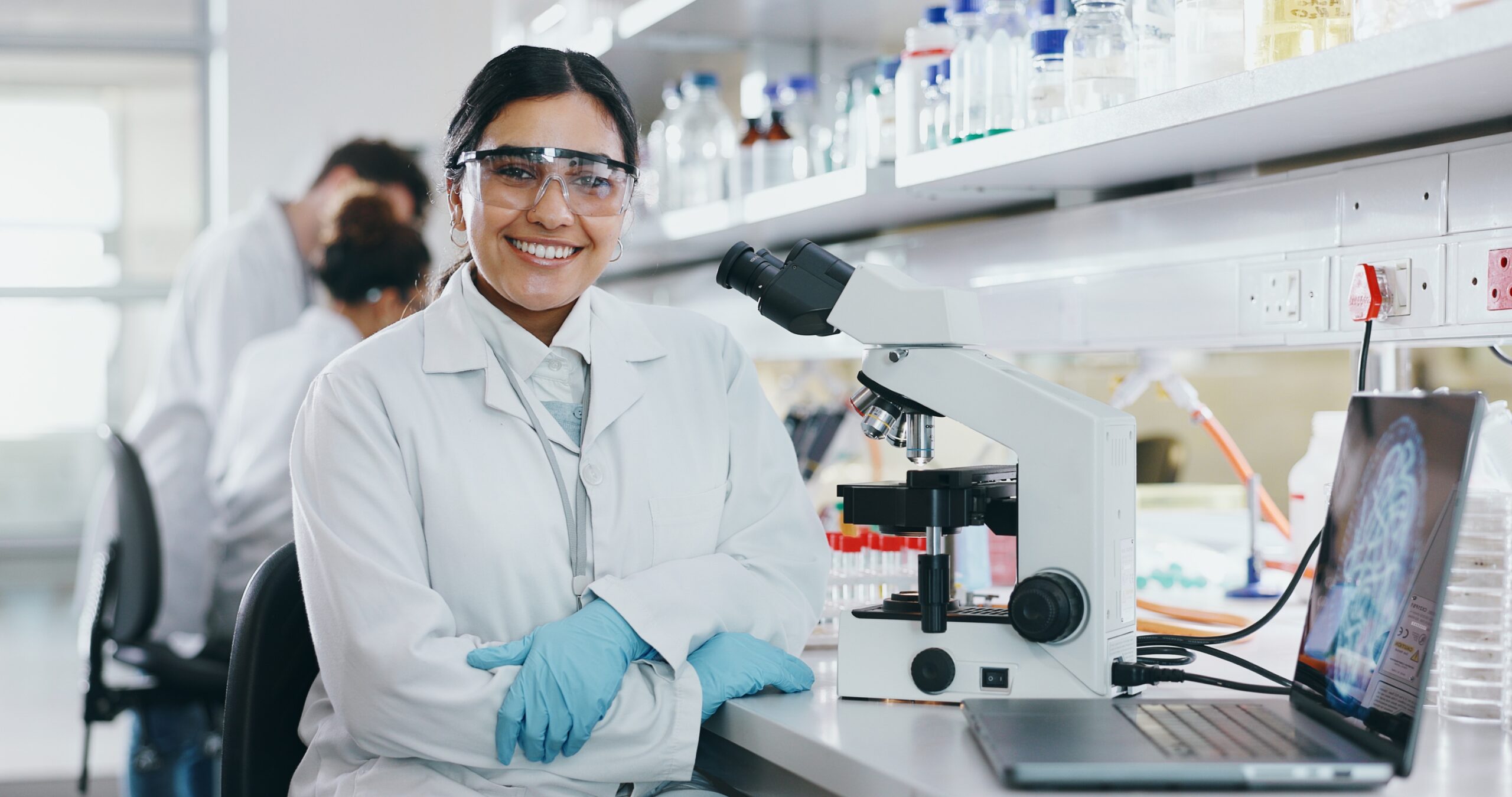A young scientist in a laboratory sits with her microscope and smiles for the camera.